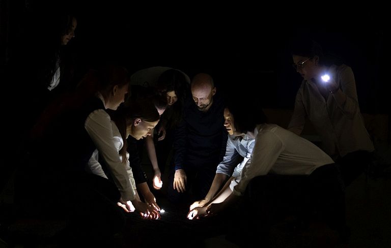 Foto aus einer Theateraufführung: Gruppe von Menschen beugt sich im Dunkeln über leuchtende Gegenstände am Boden